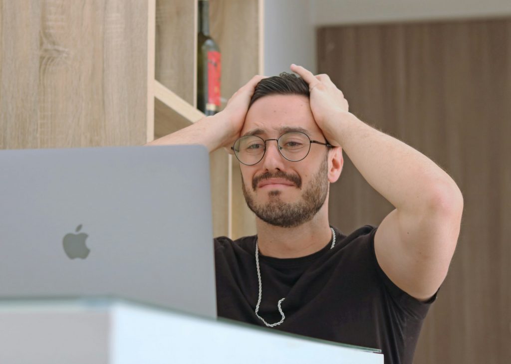 Ein dunkelhaariger Mann mit Brille der vor einem Macbook sitzt und seine Hände über dem Kopf zusammenschlägt. Zur Symbolisierung für Fehler in der Selbständigkeit