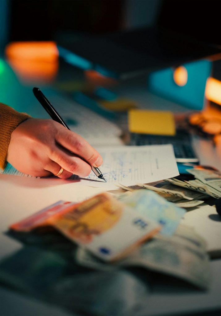 Eine schreibende Hand mit einem Ring an Finger. Neben dem Blatt, auf dem geschrieben wird liegt Bargeld.